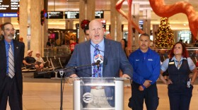 Man in a suit stands at a podium. Other officials, travelers, and a giant fiberglass and resin flamingo are behind him.