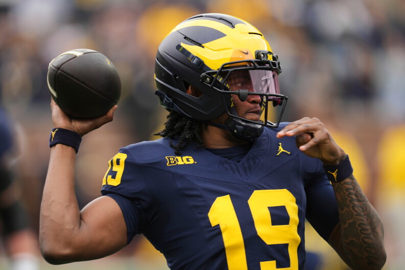 Michigan quarterback Bryce Underwood in a blue and yellow uniform prepares to throw a football during a game; stadium crowd blurred in the background.