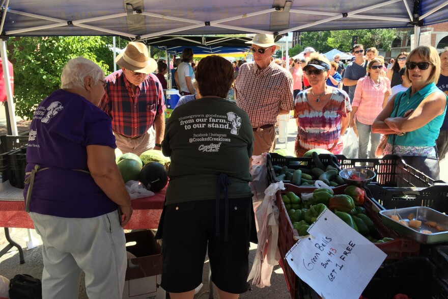 People stand around a table with melons, peppers and cucumbers.