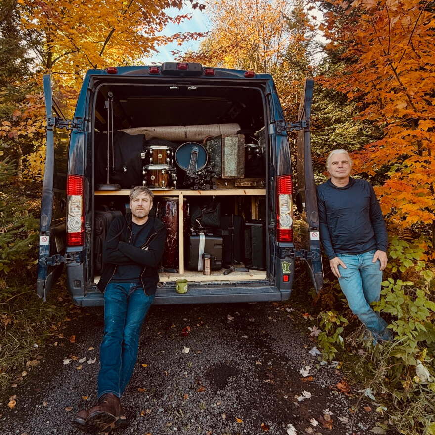 Two men posing with van that is open showing tons of musical gear in it. 