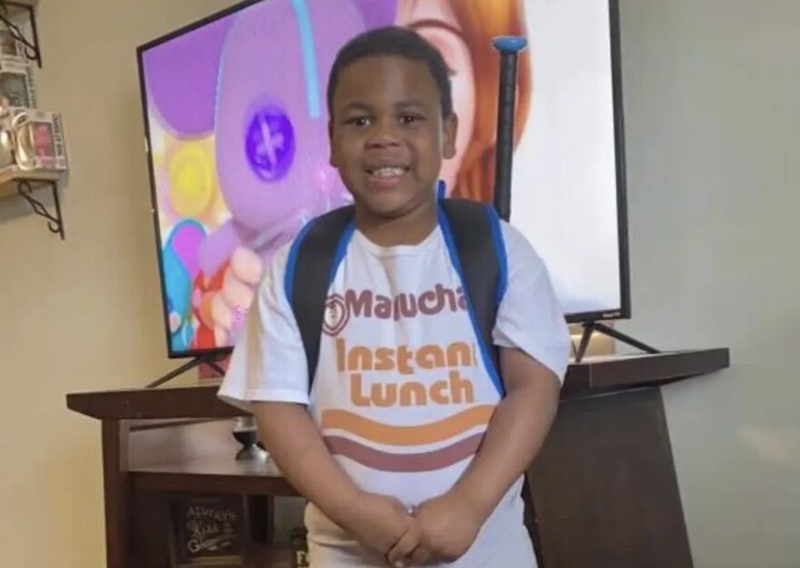  Ja'Ceon Terry, a 7-year-old child in foster care, looks at the camera and smiles. He's wearing a backpack and clasps his hands together in front of him. 