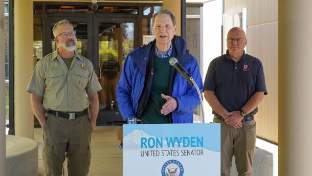 Senator Ron Wyden speaking in front of a podium with his name. Two men stand on either side. 