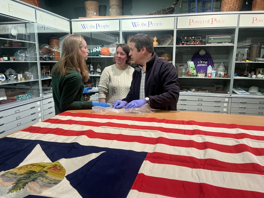 Three people wearing rubber gloves stand at a table with an American flag on it. There are cabinets of memorabilia in the background.