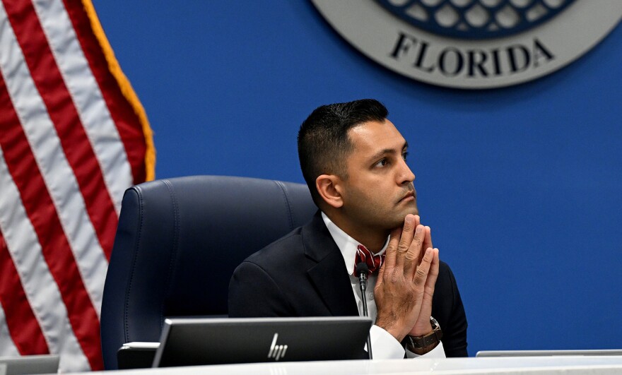 Man with short black hair sitting with his hands together on his chin in a prayer pose, looking to the right