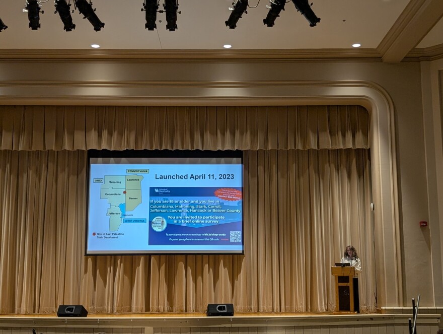 University of Kentucky researcher Erin Haynes stands behind a podium at the far right on a stage with the curtains drawn. A screen at center stage shows information about the research study.
