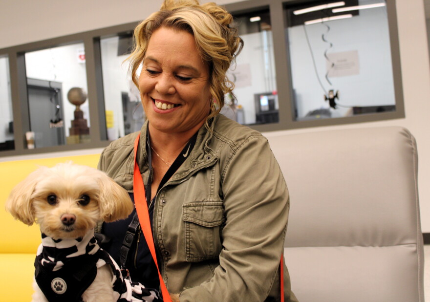 A smiling woman holds a small, light-colored dog wearing a black-and-white patterned vest.