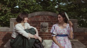 Two women sit smiling at each other on a brick fountain in early 20th century style clothing