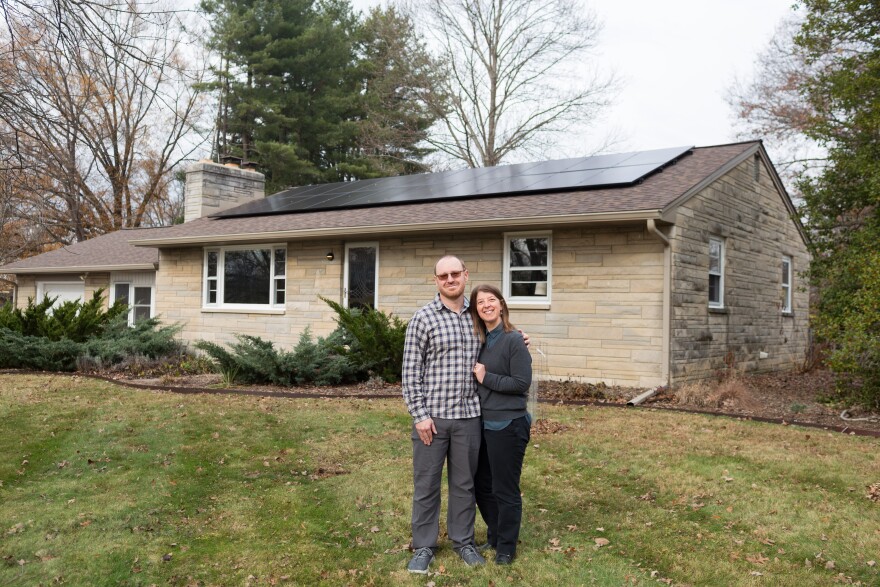 Jessica and Adam Will stand in the grass in front of their home that has solar panels on its roof. The house is stone. 