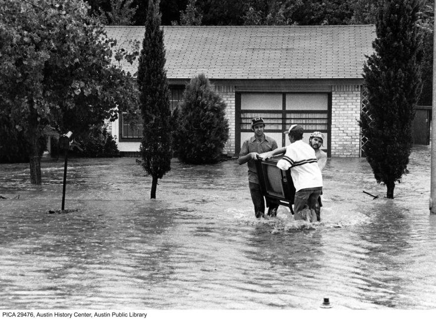 A black-and-white photograph shows three people wading through flood waters carrying a television.