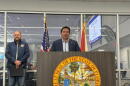Man with brown hair and a dark suit jacket standing behind a podium with a yellow state seal, in front of a glass window and a man in a blue windbreaker to the left