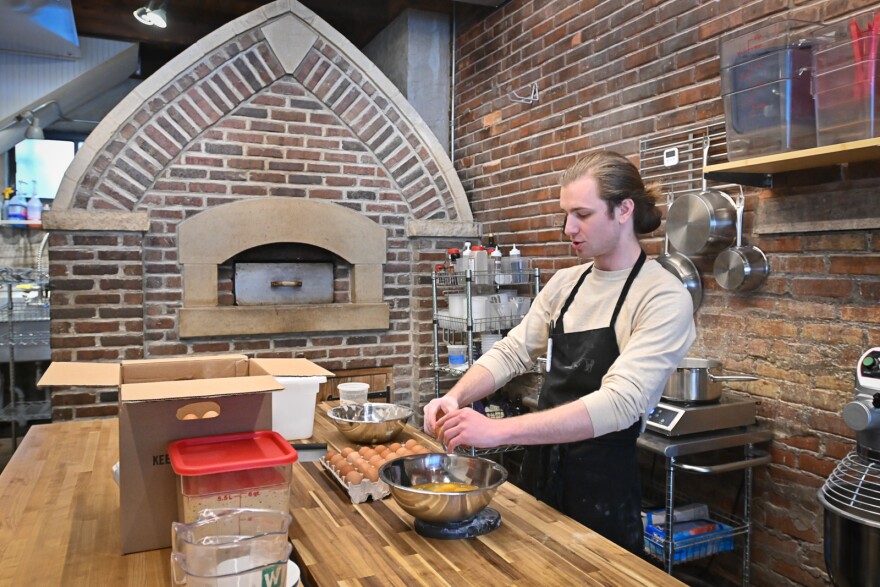 A man wearing an apron stands over a table where he is cracking eggs into a stainless steel bowl. Behind him is a brick wall and a brick oven.