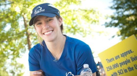 A smiling white woman in a blue t shirt and baseball cap holds a water bottle. She's sitting in a wheelchair. It's a sunny day.