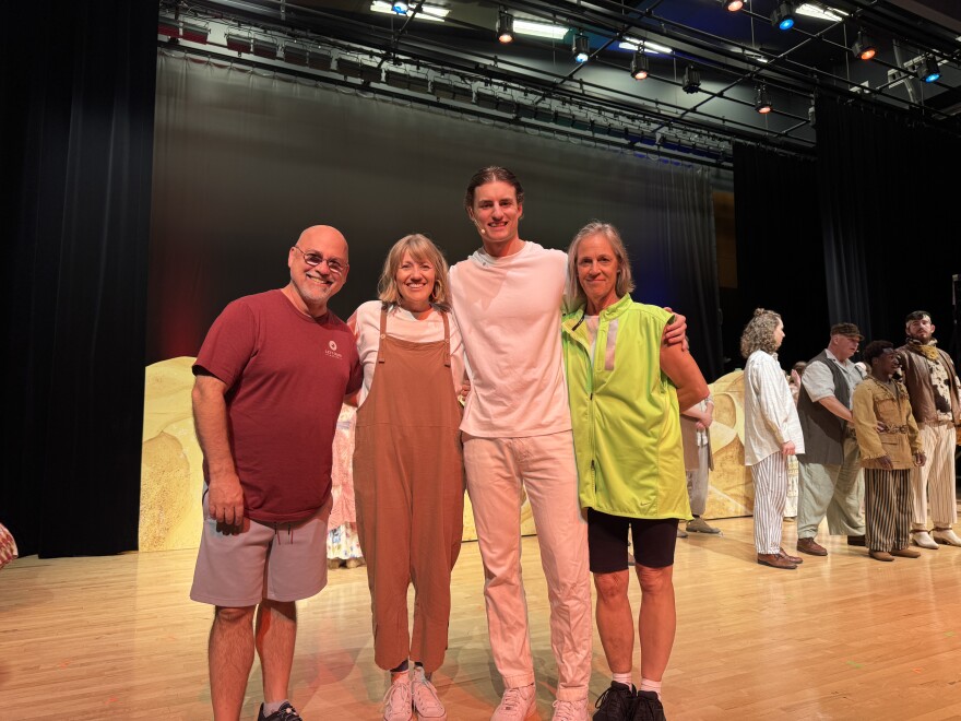 Four individuals --- two men and two women --- pose together for a picture on a stage, with cast members in the background.