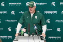 Pat Fitzgerald speaks at a podium with several microphones on it in front of a backdrop with MSU logos. He is wearing a MSU shirt and hat.