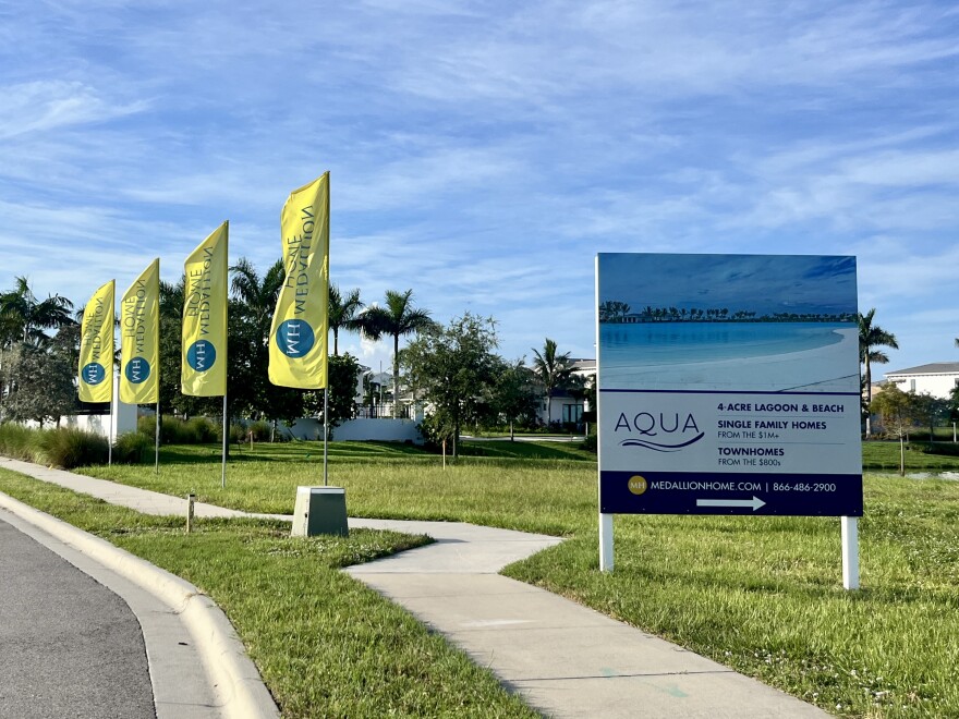 A sign outside the Aqua community. The sign reads four acre lagoon and beach, single family homes from one million plus, and townhomes from the 800s. There are four tall flags next to the sign. 