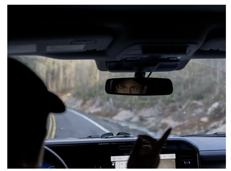 A male driver looks into a car’s rearview mirror as he points at the road outside the windshield.