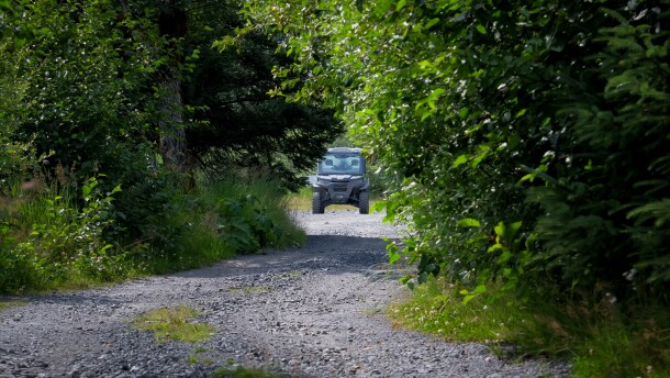 A gravel road leads up to a broken down black side-by-side, facing the camera at the center of the frame. Green trees and other foliage line the sides of trail. 