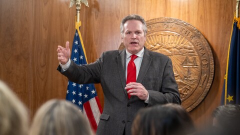 Man in wood-paneled room wearing a suit standing in front of state seal and flags