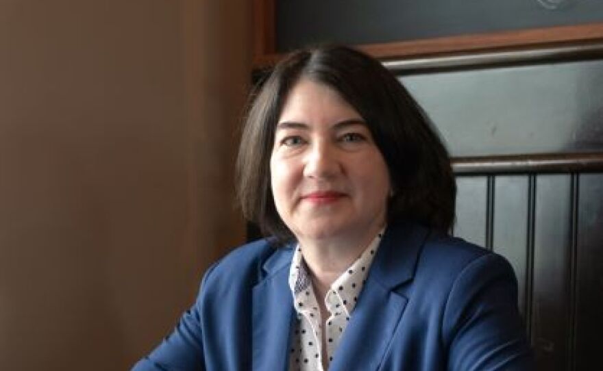 A woman with short dark hair sits in a booth wearing a navy blazer and white button-down shirt.