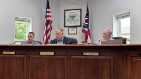 Three men sit behind a large wooden dais, with the Ohio and American flags sitting in the background.