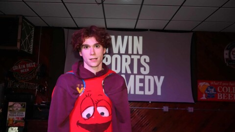A person wearing a rudimentary Red M&M costume; a poster for "Twin Ports Comedy" in the background