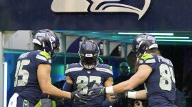 Three football players in Seahawks uniforms and helmets high five in front of a large Seahawks logo.