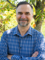 A man with brown hair and a short beard wears a plaid blue button down shirt and crosses his arms. He looks into the camera and smiles. There are trees behind him. 