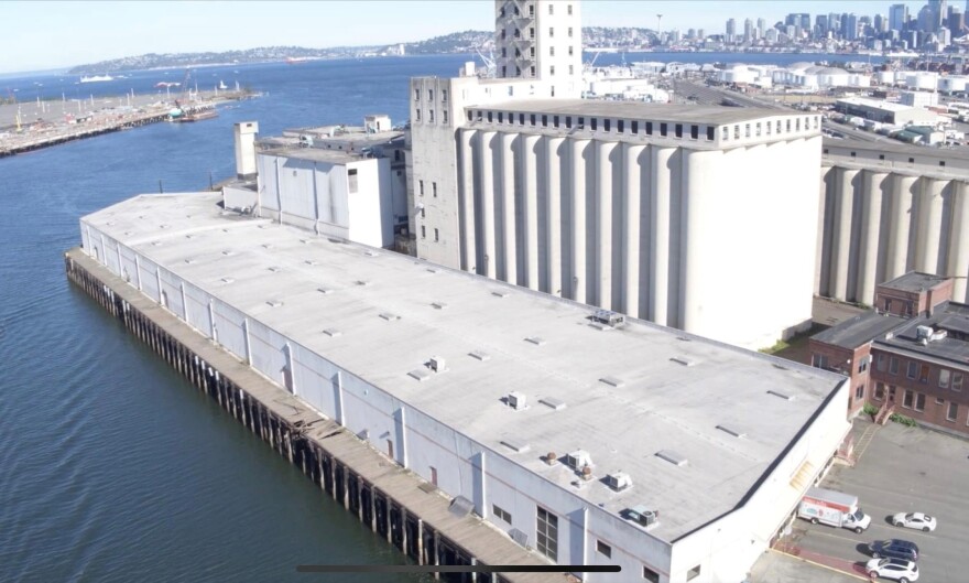 An aerial view of a white, factory-like building. The building is on a pier that juts out into water.