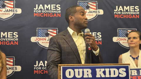 John James stands at a podium, speaking into a handheld microphone. He wears a gray plaid suit jacket and pale yellow shirt. Behind him is a campaign backdrop repeatedly bearing his name and “James for Governor Michigan.” A large sign in front reads, “OUR KIDS DESERVE BETTER.”