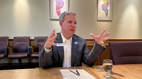 A middle aged man with gray hair sits at a conference table. He is talking and gesturing with his hands. 