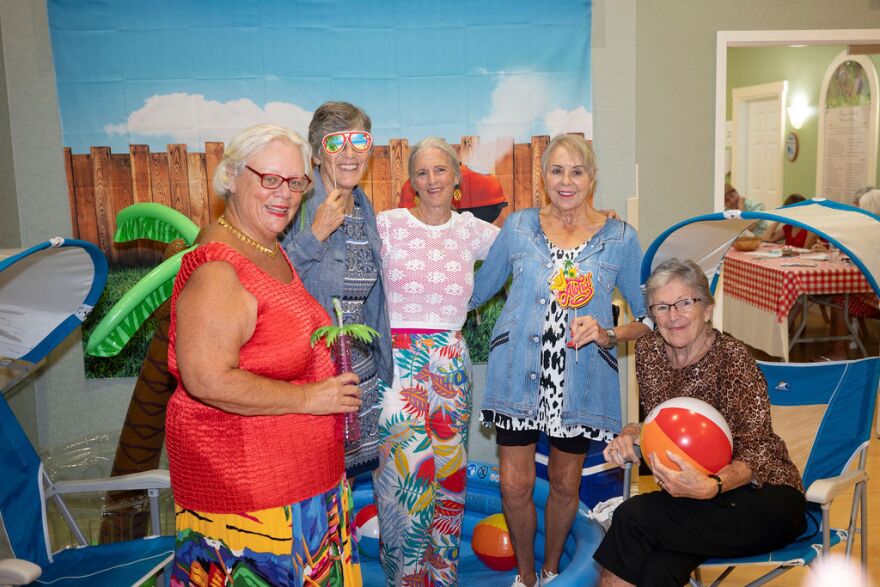 Alt text:
"Five women pose together at a beach-themed indoor event, standing and sitting in front of a backdrop of a wooden fence and blue sky mural. They wear colorful summer outfits, including a red tank top, denim jacket, and leopard-print top, with one woman wearing oversized sunglasses. A red-and-white beach ball and inflatable palm tree decorations are visible nearby, along with blue beach chairs in the foreground.