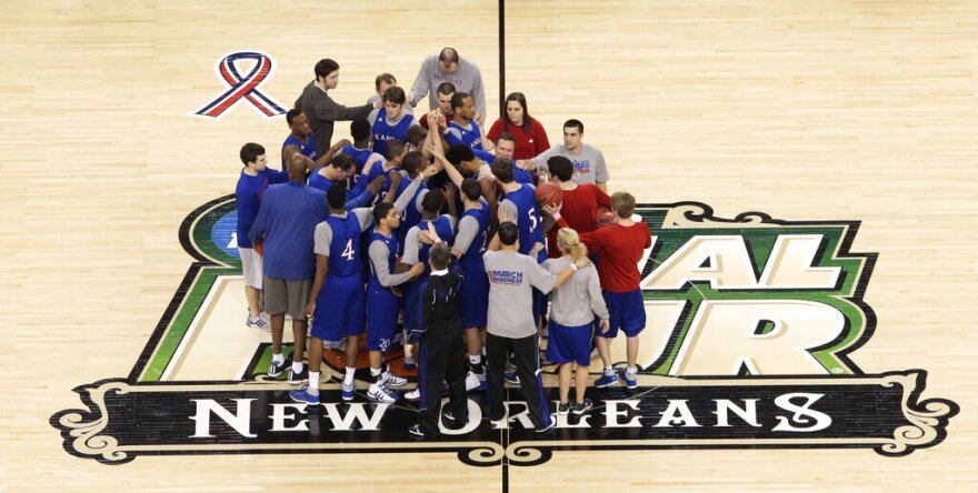 A group of basketball players and coaches gather in a huddle with arms reaching high toward the center. They are standing on a basketball court logo that reads "Final Four, New Orleans."