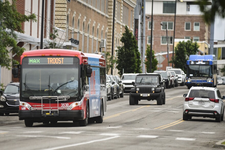 A red bus moves on a city street amidst traffic while a blue bus further back travels the same path. The red bus shows a "Max Troost" route designation. The blue bus shows a "25 Troost 83rd & Troost" designation.