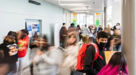 A crowd of students moves through a sunny hallway. The camera blurs their movement.