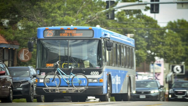 A blue transit bus moves along a busy street. On its sign designation, it reads "83rd & Troost. It appears to be moving past an intersection where other cars are just behind it. A bicycle sits on the front rack of the bus.