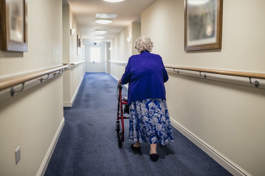 A woman with white hair walks down a blue carpeted hallway with the help of a red walker with wheels.