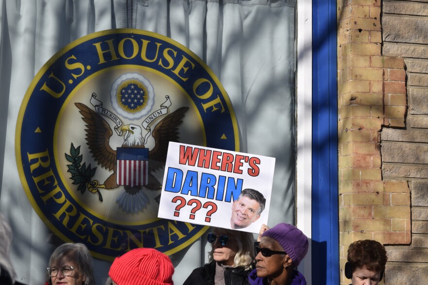 A group of protestors stand in front of a window with the U.S. House of Representatives logo on the window. A woman holds up a sign that says, "Where's Darin?" and a picture of Rep. Darin LaHood.