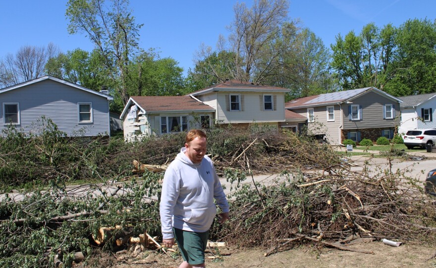 Man in a gray hoodie and green shorts walks away from a line of damaged tree limbs and other debris lined up along a residential sidewalk