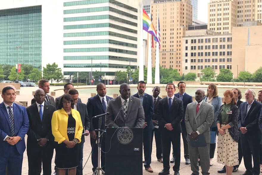 A group of people in professional attire gather outside, and a man in a suit and tie speaks at a podium.
