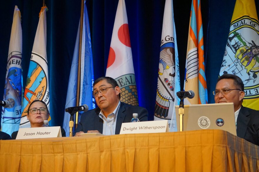 Three people sit  at a table in front of a variety of flags. Two name tags read "Jason Hauter" and "Dwight Witherspoon". There is a microphone in front of each person. 