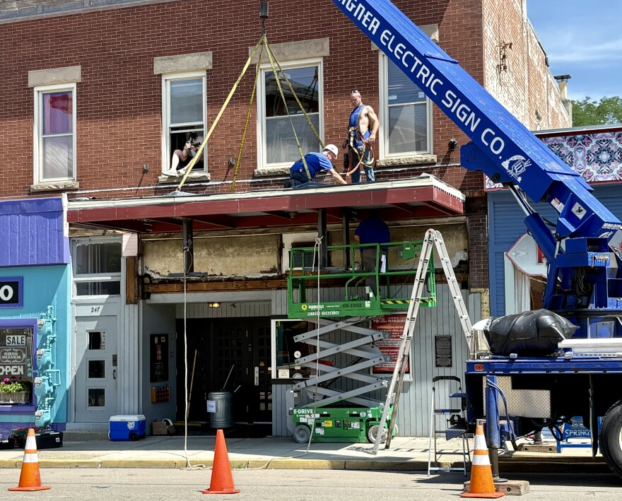 Traffic cones and barriers block off the sidewalk and part of the road outside the theater. A crane helps support the base structure for the new marquee while two workers secure it from the top.