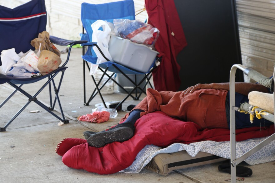 A man in orange pants sleeps on a red sleeping bag next to camping chairs and trash and clothing under an overpass.