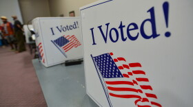 Cardboard voting dividers are shown sitting on tables with the words "I Voted!" printed in red, white and blue. In the background, some people can be seen standing.
