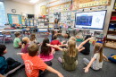 Elementary students are seated on the floor of a classroom facing their teacher, and are flapping their arms like they are flying.