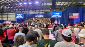 Many supports stand in a large venue with screens saying "Iowa Welcomes President Donald J Trump." 