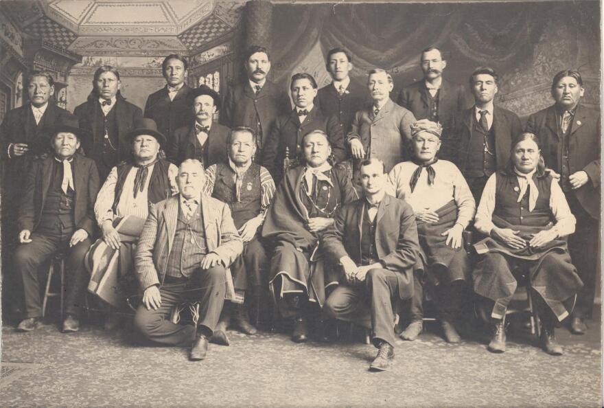 Photograph of Osage Delegation to Washington, DC in regard to losing land. Left to right: Two men kneeling in the front: Tom Mosier, Interpreter, Oscar A. Mitscher, U.S. Indian Agent. Seated row: — Brave, Heh-scah-moie, Ne-kah-wah-she-tun-kah, Back Dog, Shun-kah-mo-lah, Peter C. Bigheart. Next row (standing): Charles N. Prudom (diminutive man wearing a hat), Arthur Bonnicastle, W. S. Mathews, James Bigheart, and Wah-sho-she--(first name?) Lawrence. Top row (standing): Frank Corndropper, Bacon Rind. Fred Lookout, W. T. Leahy, Eves Tall Chief, and Charles Brown.