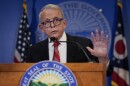 a man in blue framed glasses and a white shirt, red tie and black suit jacket talks at a podium. behind him are the american flag, ohio state flag and ohio state seal