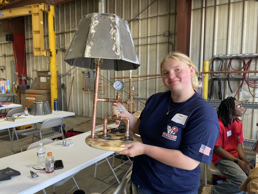 Ava Ayres poses with the project she's working on at the Heavy Metal Summer Experience. Students welded the lampshade together from pieces of sheet metal, soldered copper pipes to make the base, and ran electrical wires to the lightbulb.