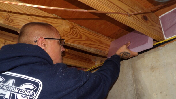 An over-the-shoulder image of a bald man in a navy blue hoodie placing a piece of purple foam insulation in between the beams of a ceiling. 