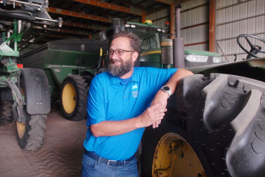 A man wearing glasses, a blue polo and jeans leans against the wheel of a green tractor in a barn. 